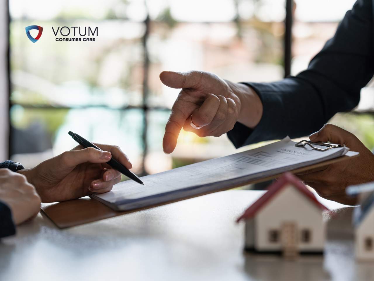 <br />
Zbliżenie na dokument umowy, przy którym dłoń osoby w garniturze wskazuje na konkretne zapisy, podczas gdy inna dłoń trzyma długopis, a w dolnym prawym rogu widoczne są miniaturowe modele domów