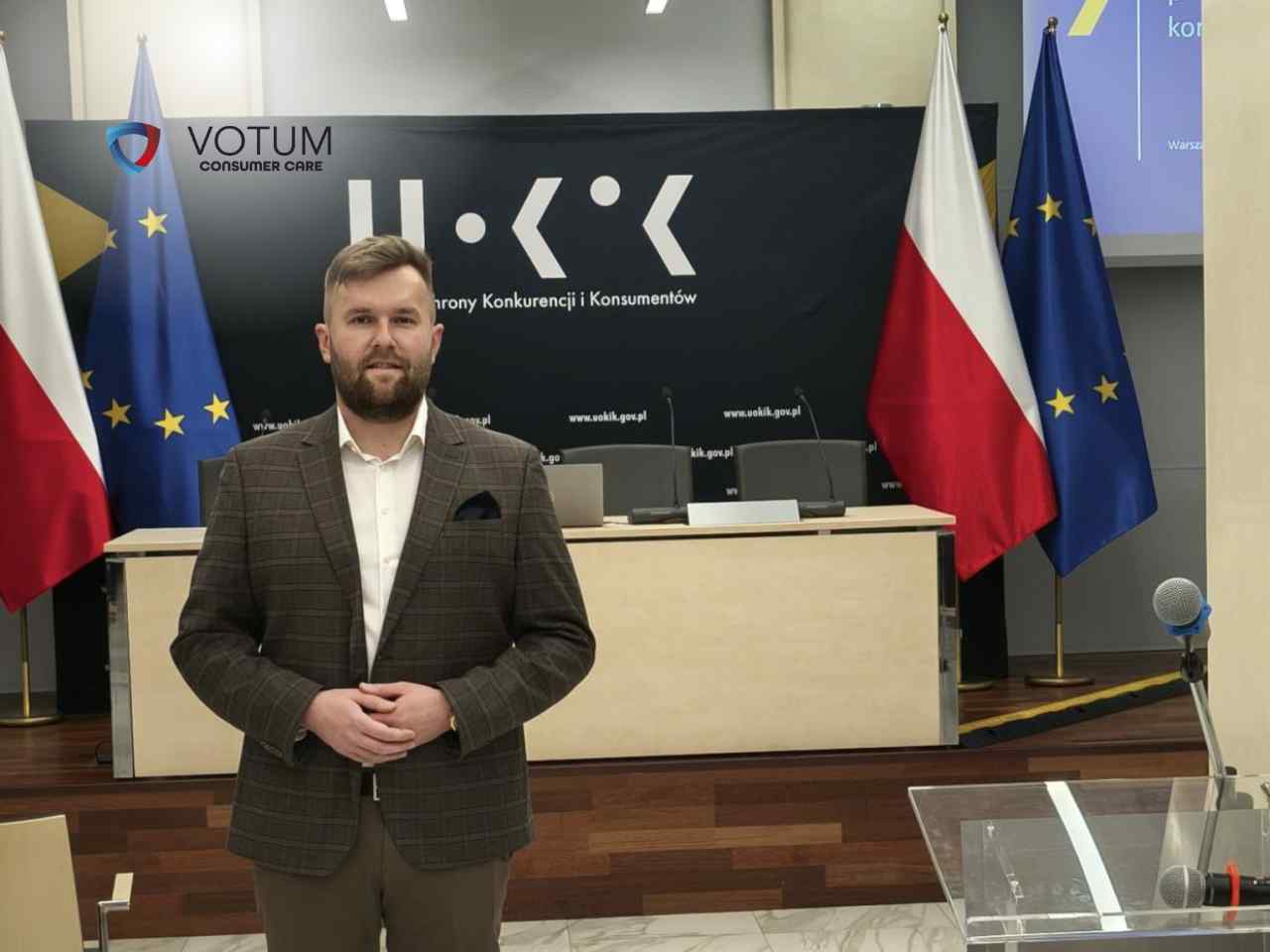 Grzegorz Bartoszek, Dyrektor Departamentu Spraw Bankowych, stoi na tle flag Polski i Unii Europejskiej oraz ścianki z logo UOKiK