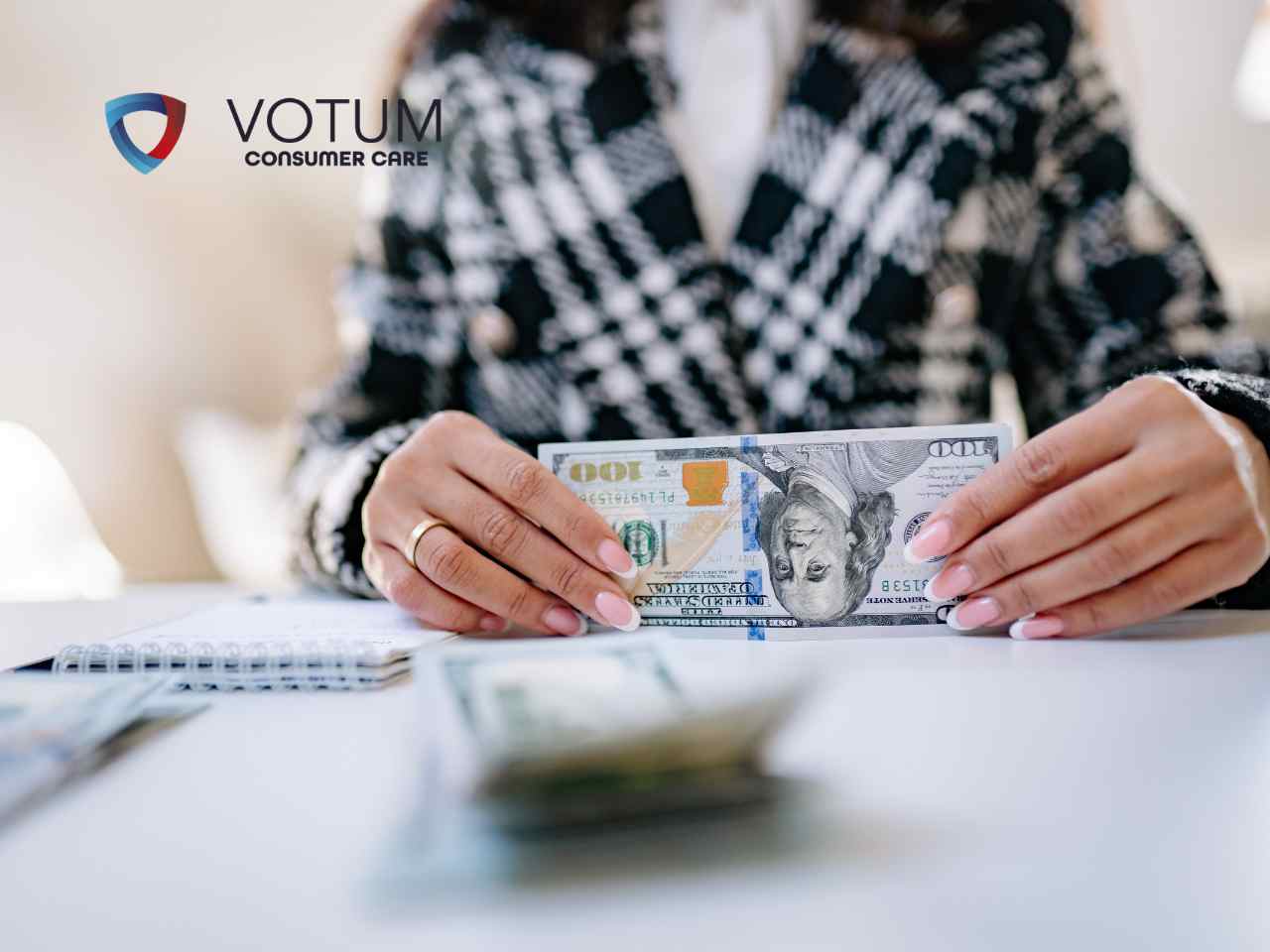 umowy-kredytow-waloryzowane-do-USD Kobieta w marynarce w kratę trzyma w dłoniach banknot o nominale 100 dolarów amerykańskich nad białym biurkiem.