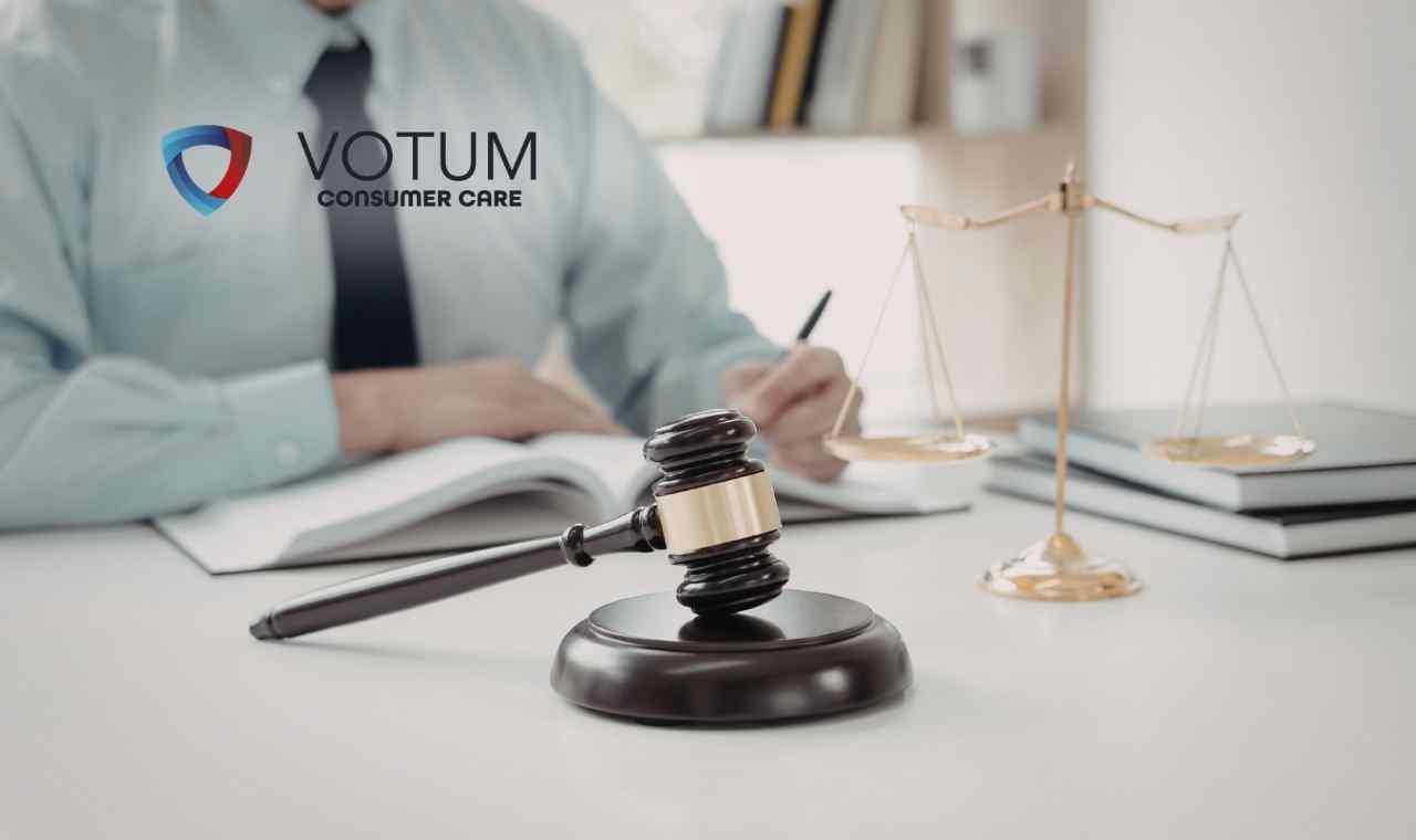 Na pierwszym planie widoczny jest ciemny, drewniany młotek sędziowski na podstawce; w tle, na rozmytym planie, znajduje się waga szalkowa oraz osoba w błękitnej koszuli z krawatem, która sporządza notatki w dokumentach.