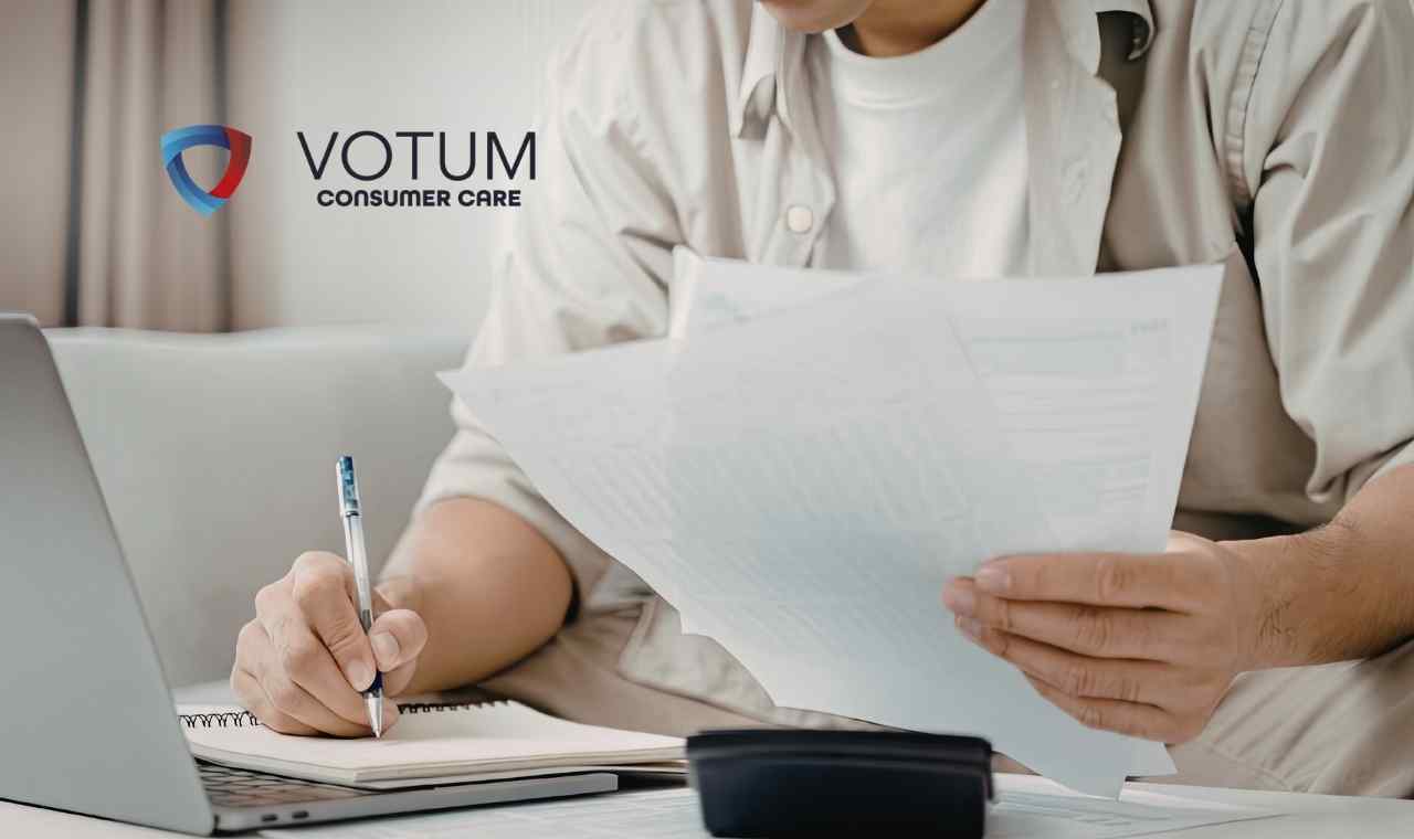 Zbliżenie na dłonie osoby siedzącej przy biurku, która analizuje dokumenty kredytowe. Jedna ręka trzyma arkusze papieru, a druga robi notatki w zeszycie. Na biurku widoczny jest również fragment laptopa oraz kalkulator.