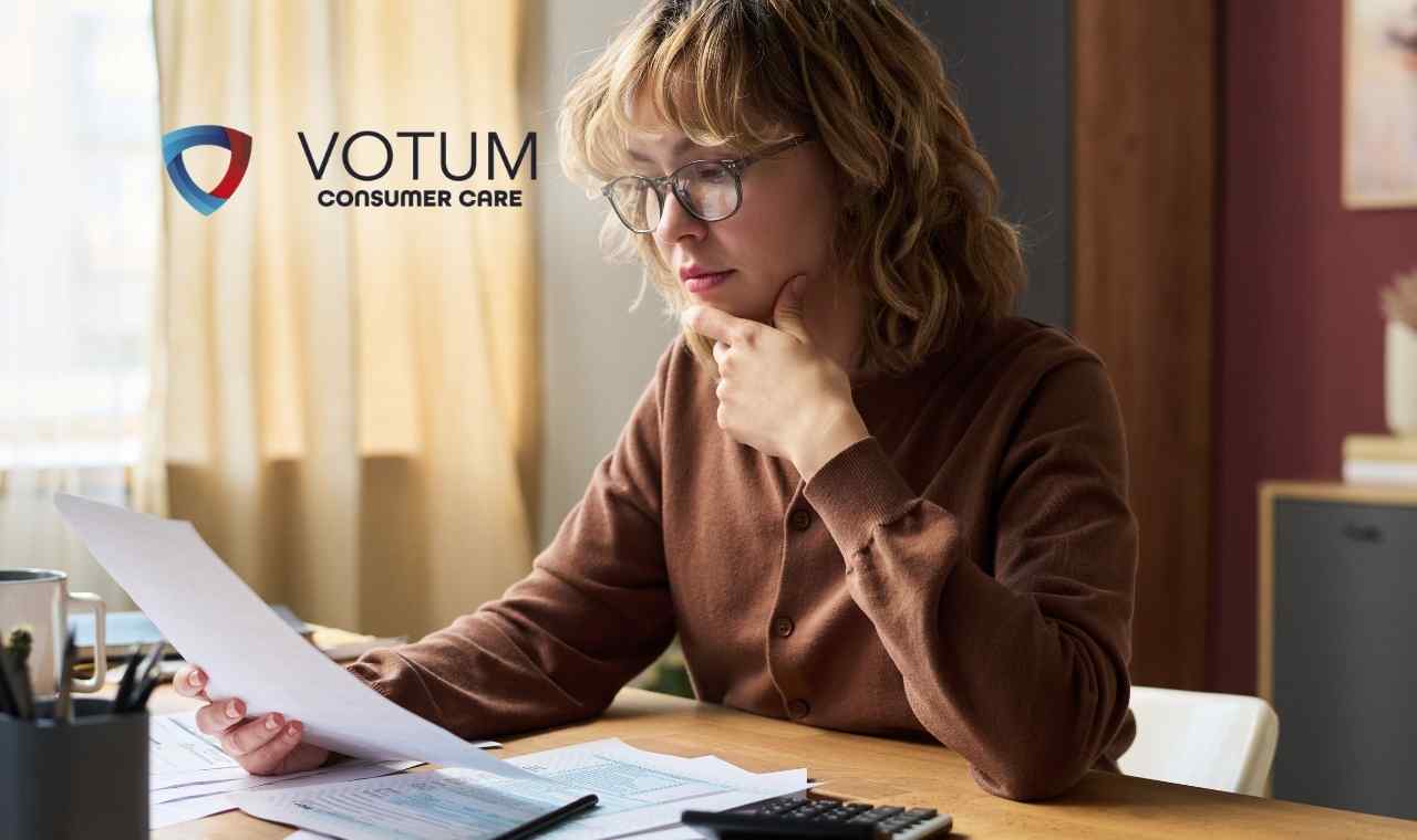 Kobieta w okularach i brązowym kardiganie siedzi przy biurku, trzymając dłoń na podbródku i z wyraźnym skupieniem analizując trzymaną w ręku kartkę papieru. Przed nią leżą inne dokumenty oraz kalkulator.