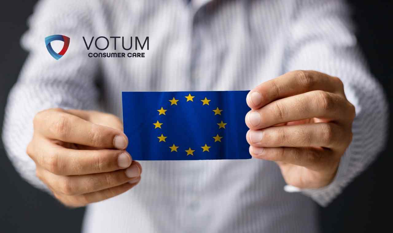 Zbliżenie na dłonie osoby w jasnej, prążkowanej koszuli, która oburącz trzyma małą, prostokątną kartę z flagą Unii Europejskiej.