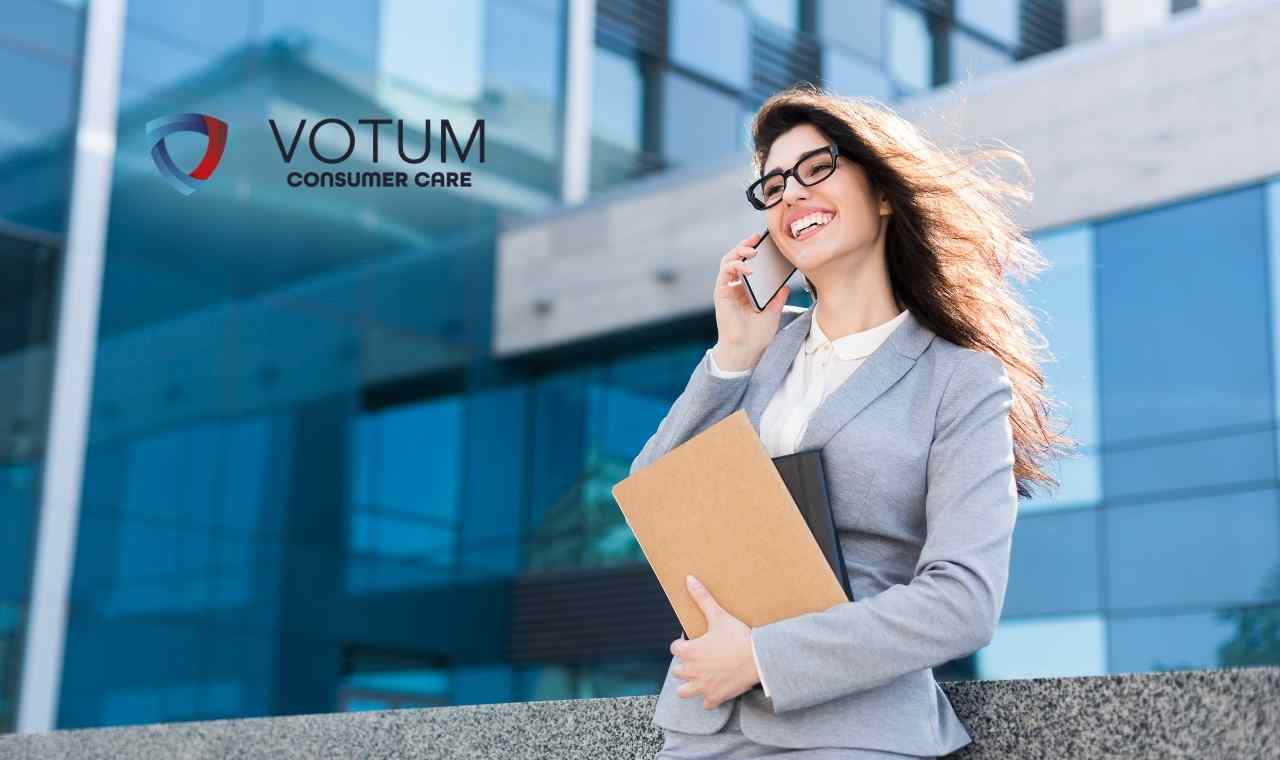 Uśmiechnięta kobieta w szarym żakiecie i okularach rozmawia przez telefon, trzymając pod pachą brązową teczkę z dokumentami. W tle widoczny jest nowoczesny biurowiec o przeszklonej fasadzie. W lewym górnym rogu znajduje się logotyp Votum Consumer Care.