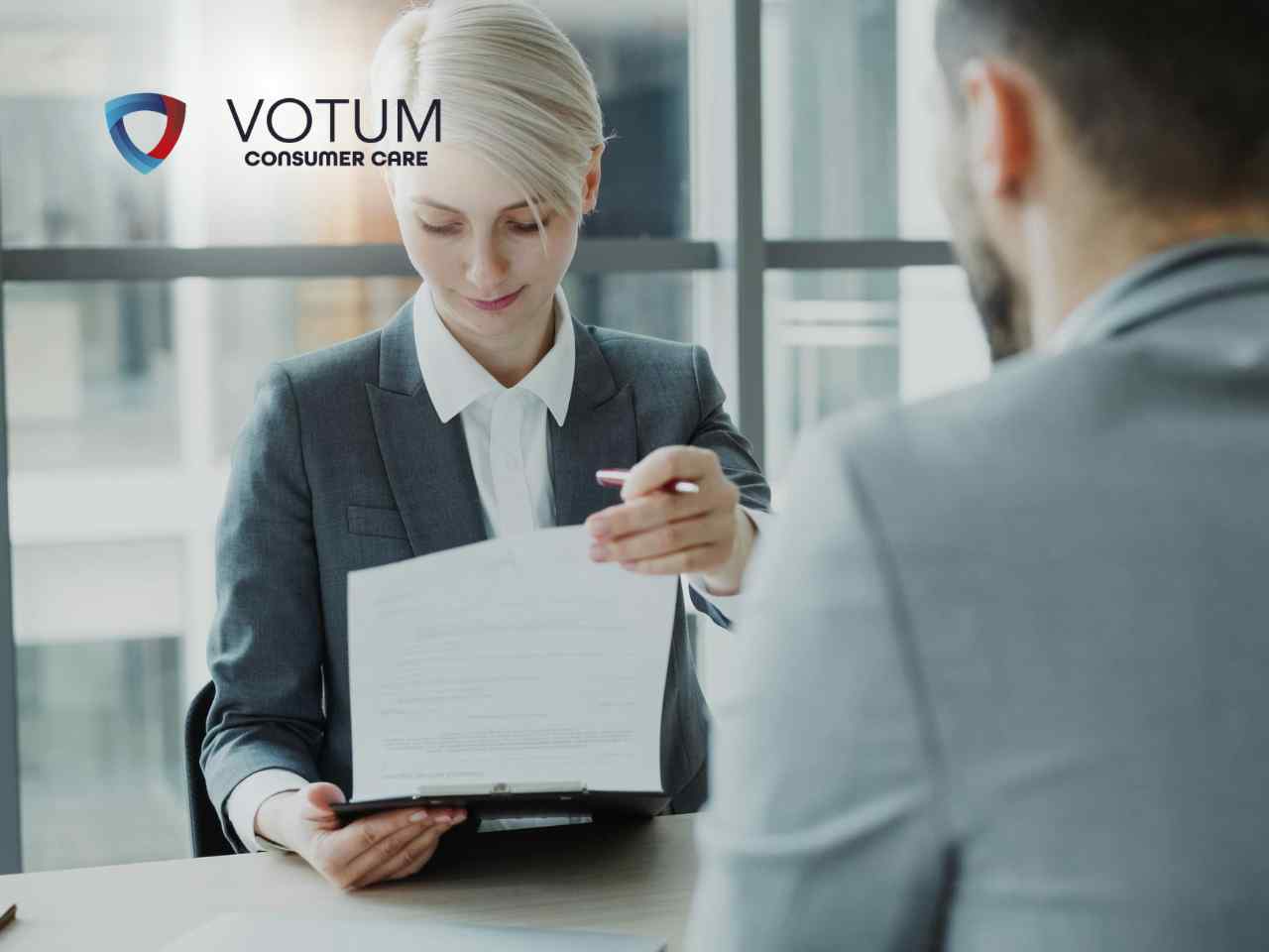 Kobieta w ciemnoszarej marynarce (prawniczka) siedzi przy biurku naprzeciwko klienta (widocznego od tyłu). Z lekkim uśmiechem i profesjonalnym skupieniem prezentuje klientowi dokument umieszczony w czarnym folderze, wskazując długopisem na konkretny zapis.