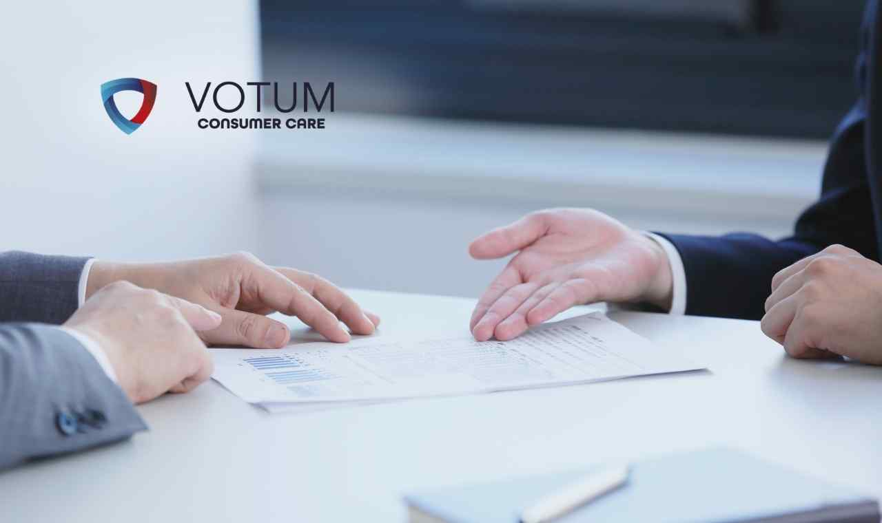 Dwie osoby w garniturach siedzą przy białym stole; jedna z nich wykonuje gest dłonią, wyjaśniając treść leżącego przed nimi dokumentu z wykresami kolumnowymi.