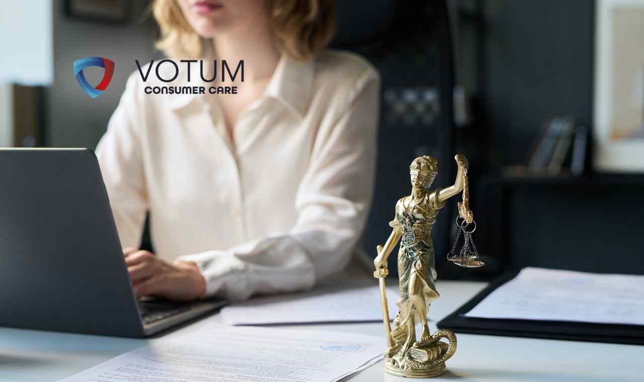 Na pierwszym planie złota figurka Temidy, bogini sprawiedliwości, stojąca na biurku obok dokumentów; w tle widać rozmytą sylwetkę kobiety pracującej przy laptopie.