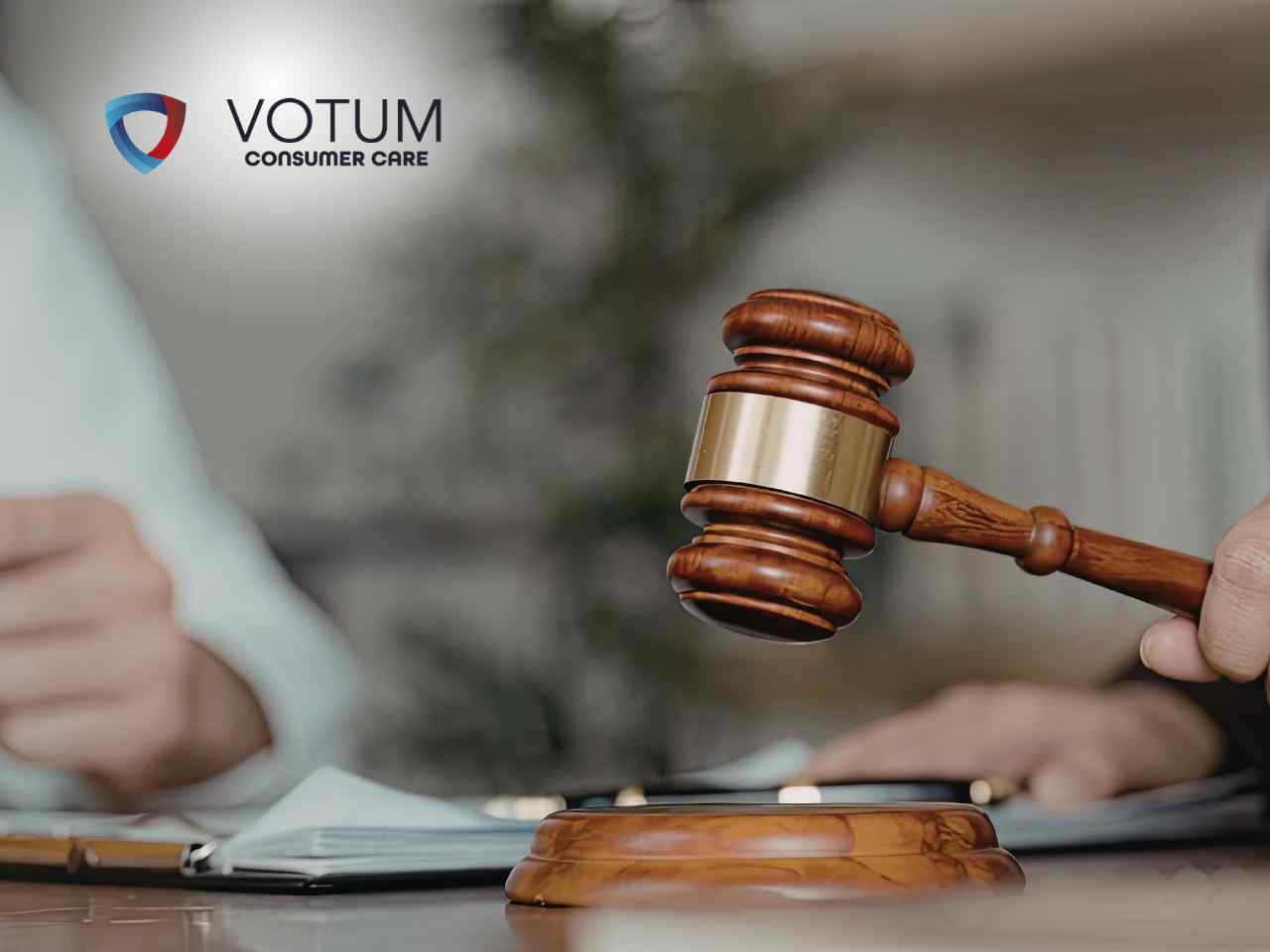 Zbliżenie na dłoń sędziego trzymającą drewniany młotek tuż przed uderzeniem w podstawkę; w tle, na rozmytym planie, widoczna jest postać pracująca przy dokumentach.