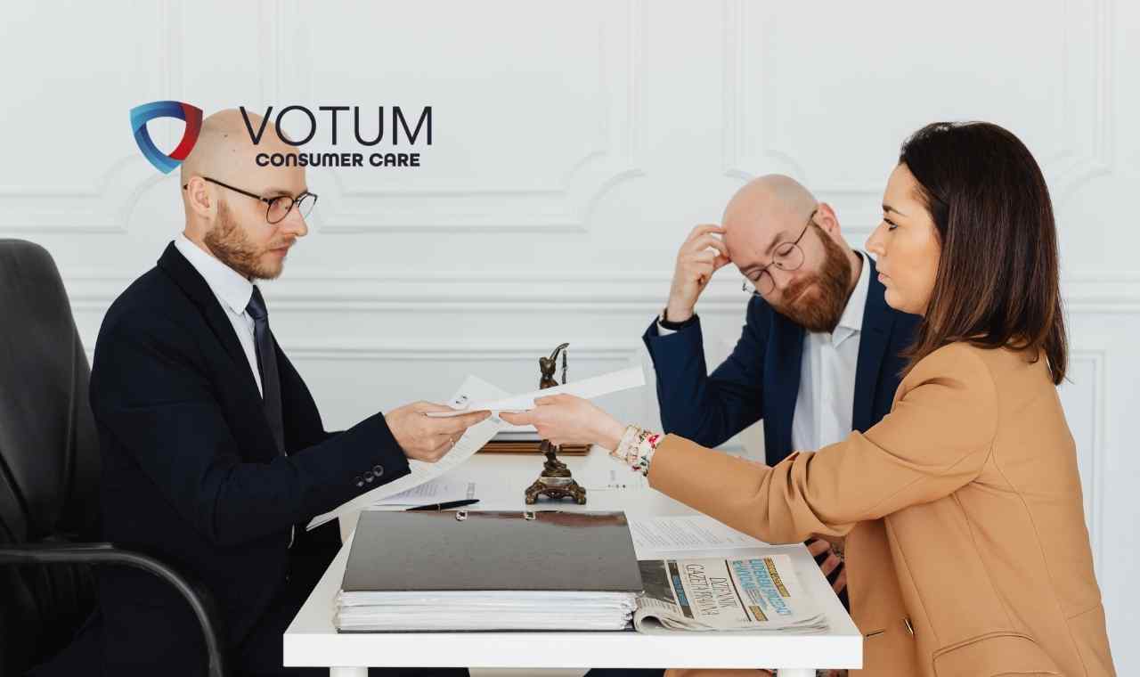 Scena w biurze lub kancelarii; po lewej stronie mężczyzna w ciemnym garniturze i okularach przekazuje dokument kobiecie w beżowej marynarce. W tle widoczny jest drugi mężczyzna, który z niedowierzaniem trzyma się za głowę. Na stole leżą teczki z aktami oraz gazeta.