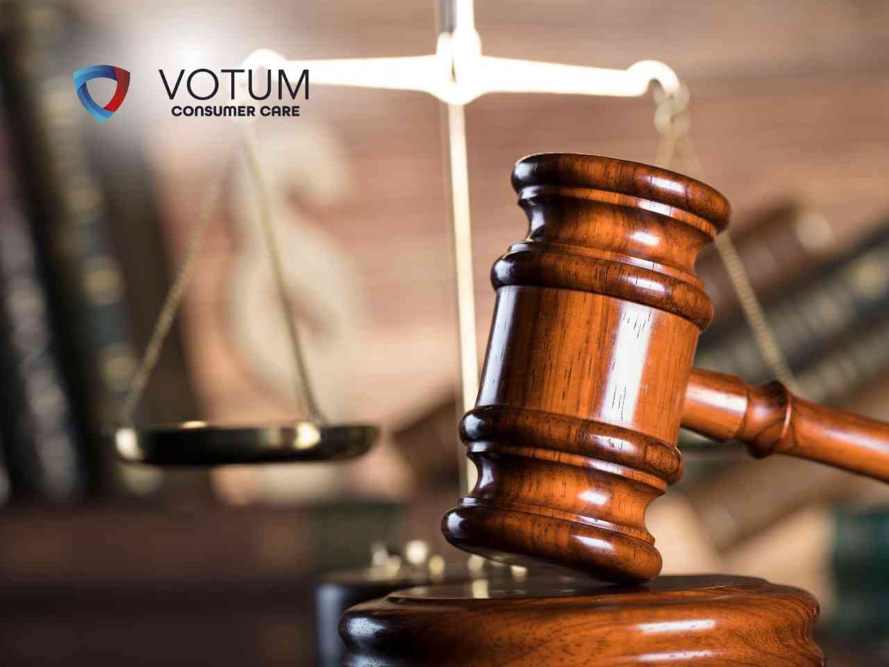 Prawnik w okularach rozmawia przy biurku z parą klientów, tłumacząc zawiłości prawne. Na stole widoczna jest statuetka Temidy oraz otwarte księgi prawnicze. W lewym górnym rogu znajduje się logotyp Votum Consumer Care.