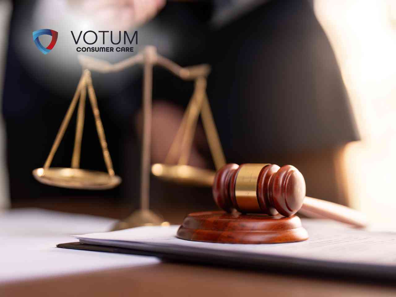 Prawnik w garniturze prezentuje dokument do podpisu klientom siedzącym przy biurku, na którym znajdują się waga szalkowa i młotek sędziowski. W lewym górnym rogu widnieje logotyp Votum Consumer Care.