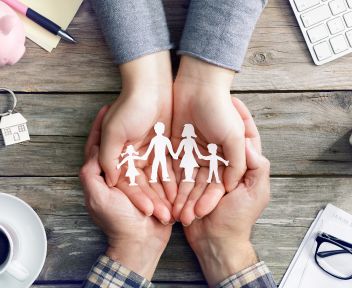 pomoc-frankowiczom-votum-consumer-care Widok z góry na drewniany stół, na którym dłonie dwóch osób tworzą ochronny gest, otaczając papierową wycinankę przedstawiającą rodzinę (rodziców i dwoje dzieci). Wokół widoczne są przedmioty codziennego użytku: filiżanka kawy, okulary, klucze do domu oraz świnka skarbonka. Zdjęcie symbolizuje bezpieczeństwo finansowe i ochronę ogniska domowego.