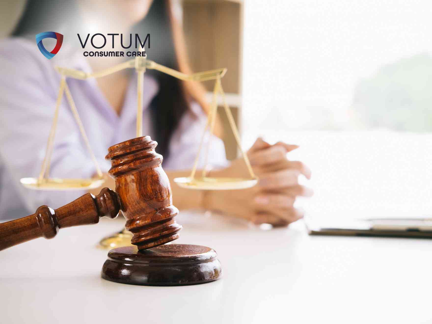 Prawnik w okularach rozmawia przy biurku z parą klientów, tłumacząc zawiłości prawne. Na stole widoczna jest statuetka Temidy oraz otwarte księgi prawnicze. W lewym górnym rogu znajduje się logotyp Votum Consumer Care.