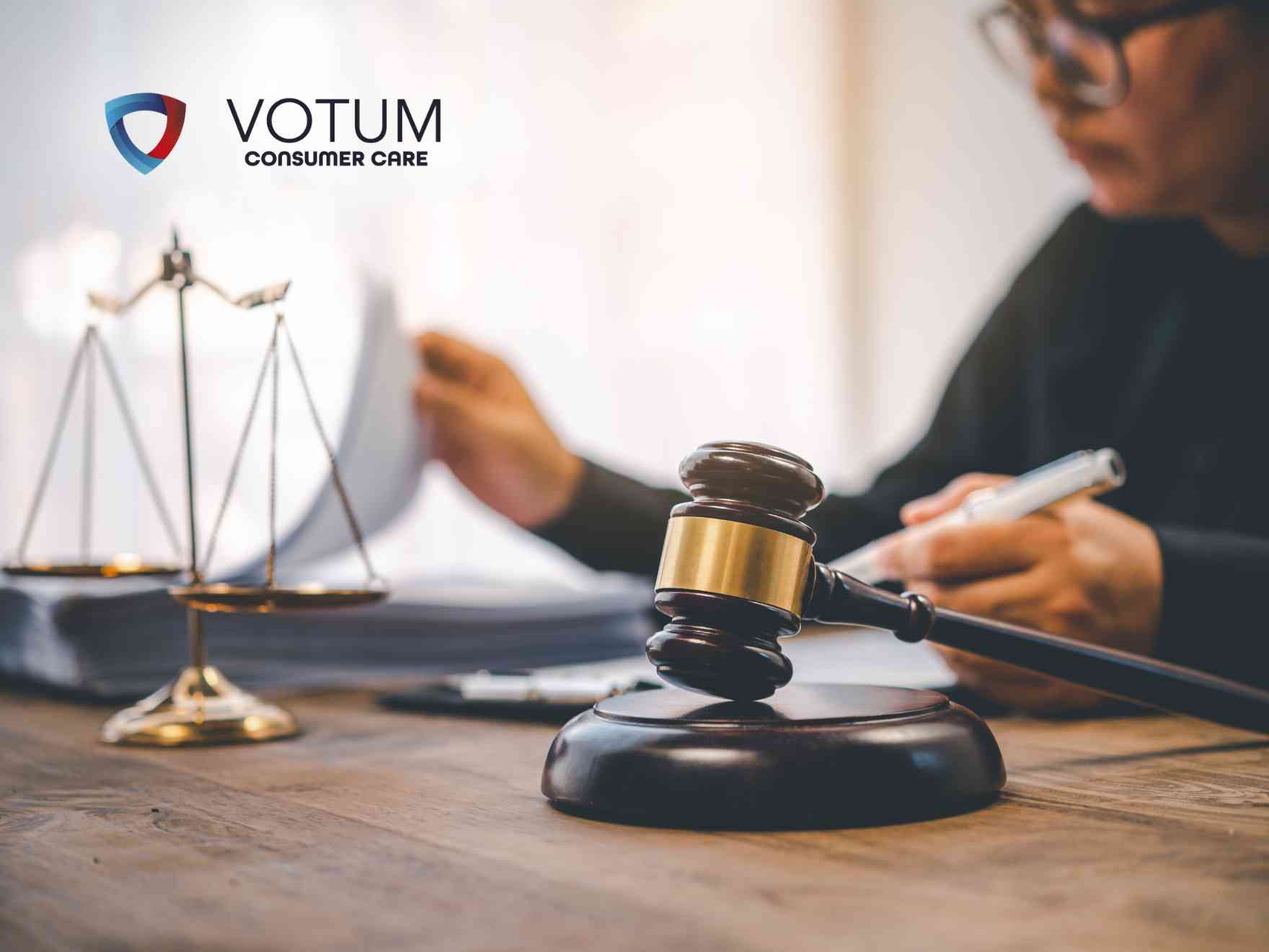 Prawnik w garniturze prezentuje dokument do podpisu klientom siedzącym przy biurku, na którym znajdują się waga szalkowa i młotek sędziowski. W lewym górnym rogu widnieje logotyp Votum Consumer Care.
