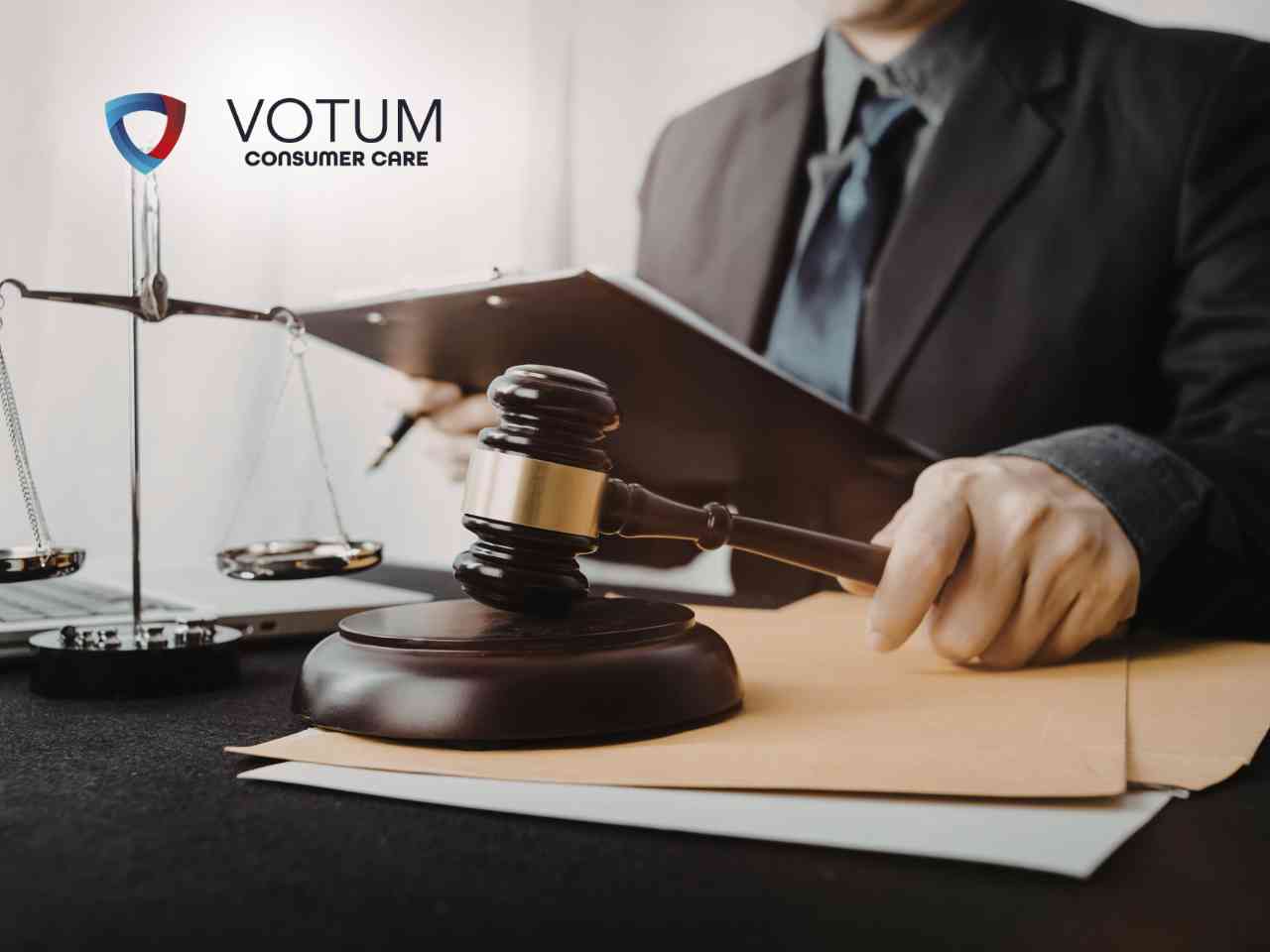 Prawnik w garniturze prezentuje dokument do podpisu klientom siedzącym przy biurku, na którym znajdują się waga szalkowa i młotek sędziowski. W lewym górnym rogu widnieje logotyp Votum Consumer Care.