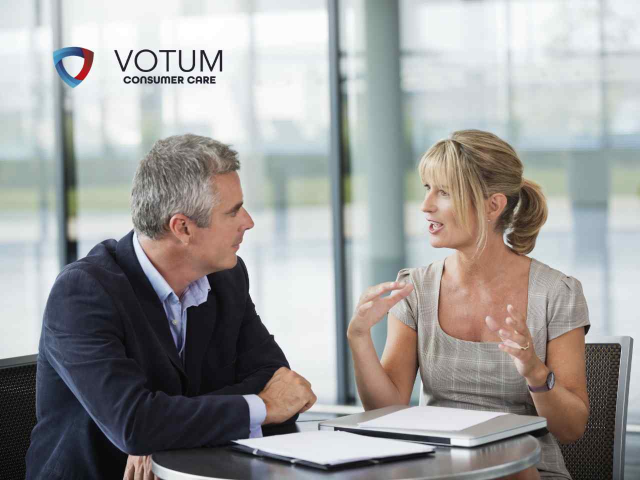 Two professionals in a meeting at a round table, discussing with documents; VOTUM CONSUMER CARE logo visible in the top left.