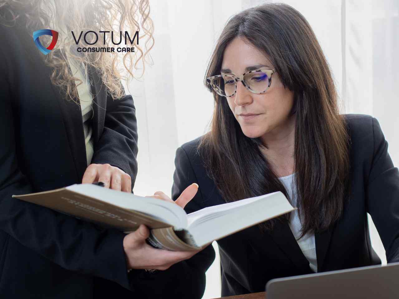 Two professional women review a document together; one points at a page while the other, with glasses, looks on, with the VOTUM Consumer Care logo in the top left corner.