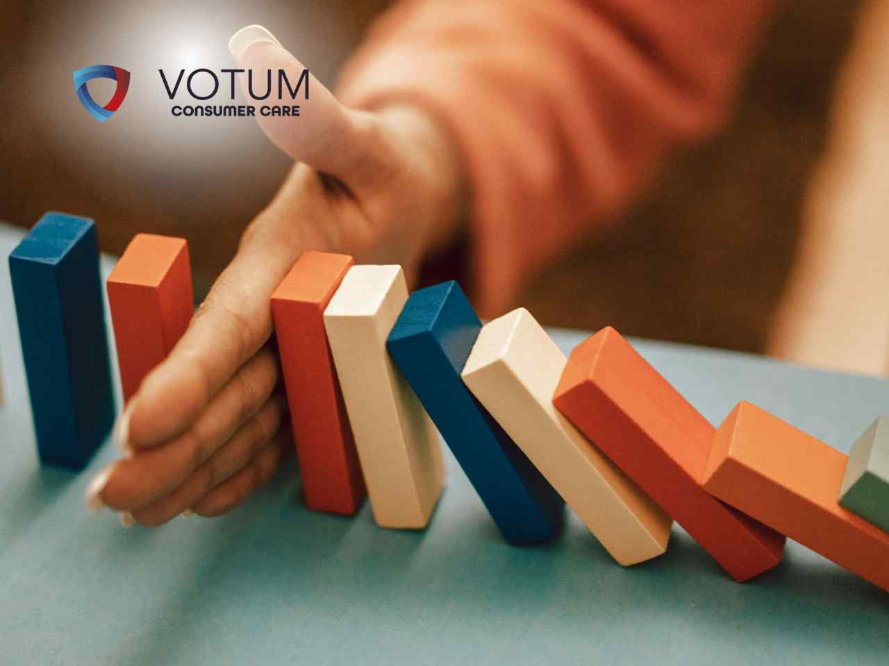 Hands gently stopping a line of colorful dominoes from falling, with VOTUM Consumer Care logo in the corner.